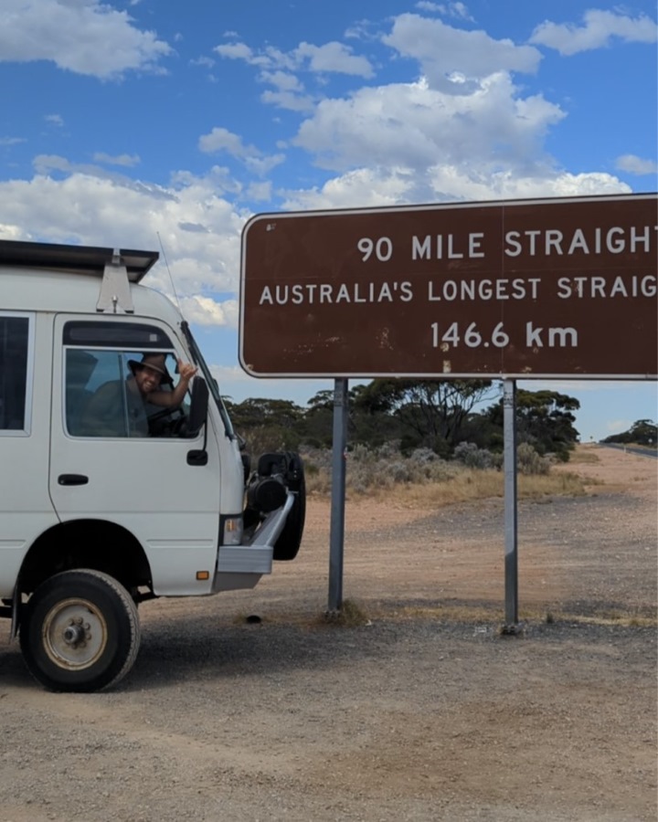 Man out the window of his caravan, waving next to the 90 mile straight sign in the Nullarbor, Western Australia