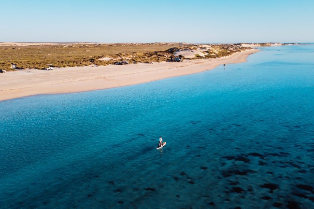 A person paddleboarding on the crystal-clear waters of Exmouth near a sandy coastline.