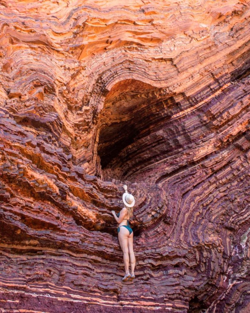 A woman in swimwear climbs the wave-like, layered rock formations in Hamersley Gorge, showcasing the vibrant reds and purples of one of the best gorges in Karijini National Park. She touches the rock wall with one hand, admiring the texture.