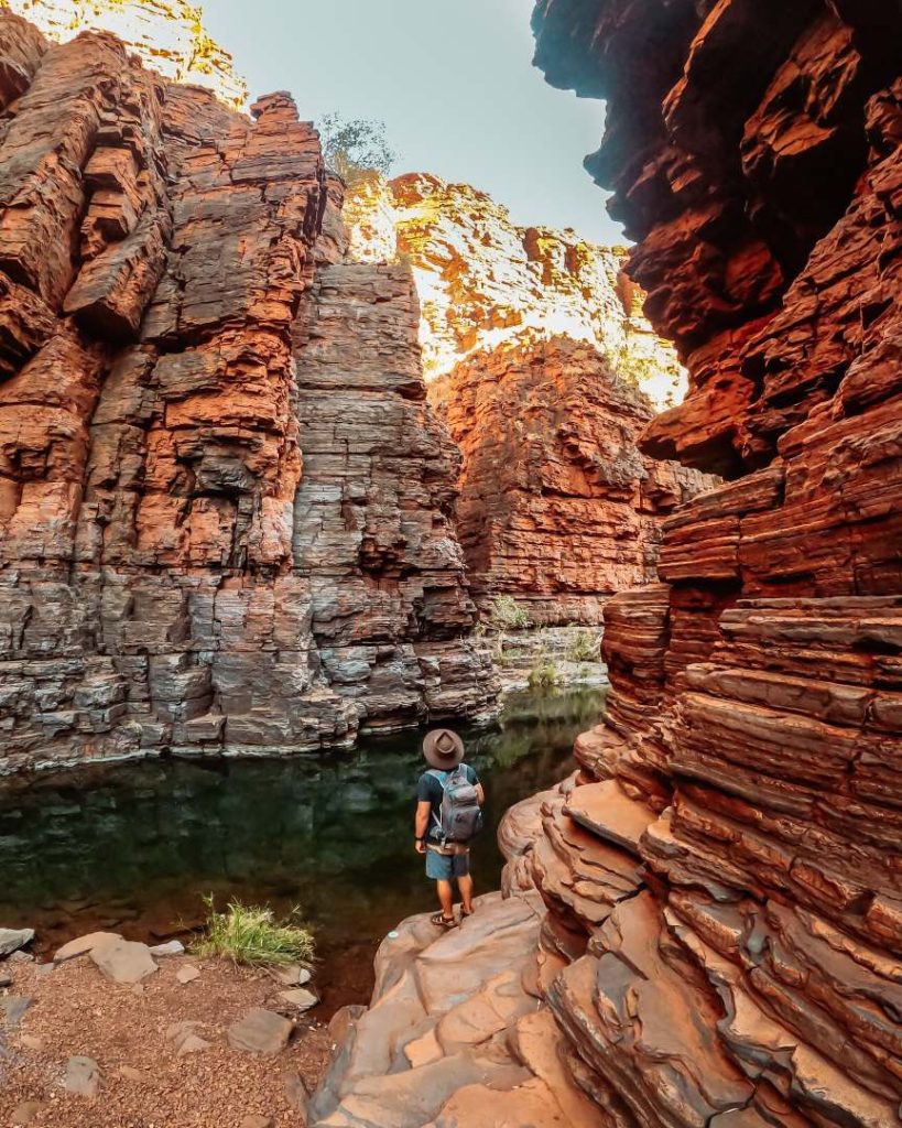 A hiker with a backpack stands at the water’s edge, dwarfed by towering orange rock walls in Joffre Gorge—one of Karijini’s highlights during the best time to visit.