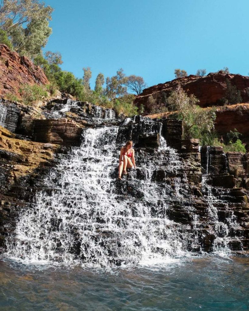 A woman sits on a rock ledge in the cascading water of Fortescue Falls, located in Dales Gorge, one of the best gorges in Karijini National Park. The waterfall flows over layered rock into a pool below.