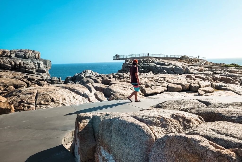 Man walking barefoot along the rocky coastline towards The Gap lookout platform in Albany, a must-see highlight on the Perth to Albany road trip.