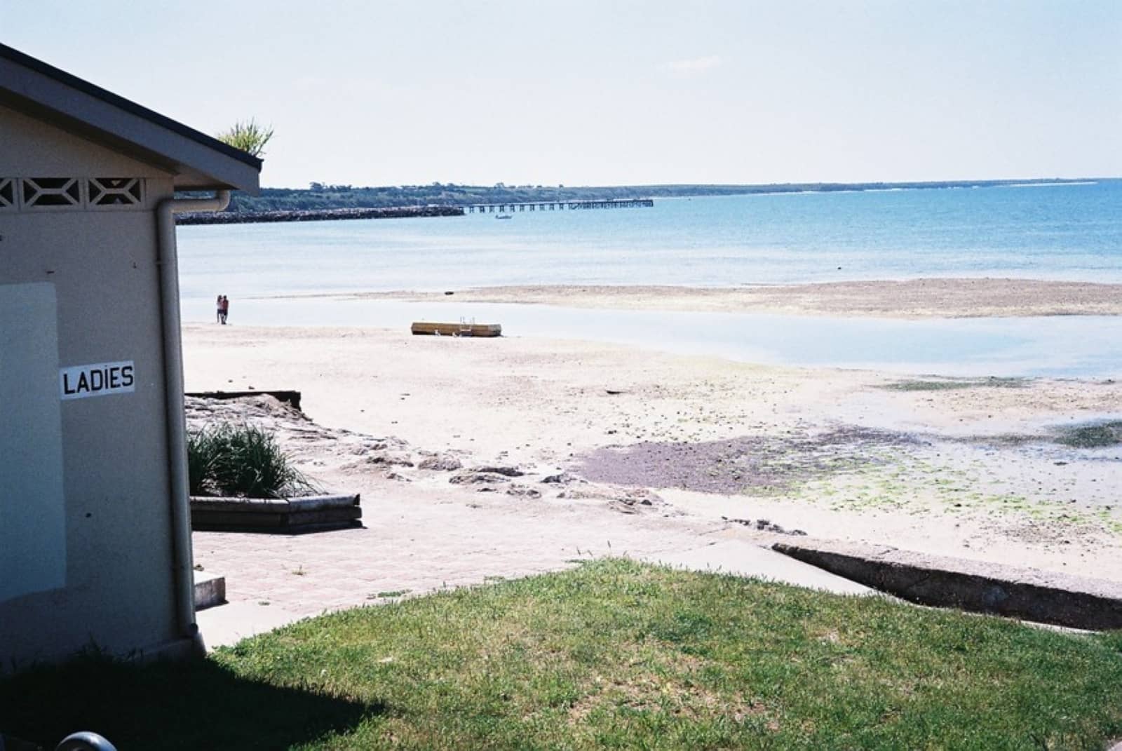 A sandy beach with calm water seen next to a public restroom building marked “LADIES,” with a couple walking in the distance during a Yorke Peninsula road trip.