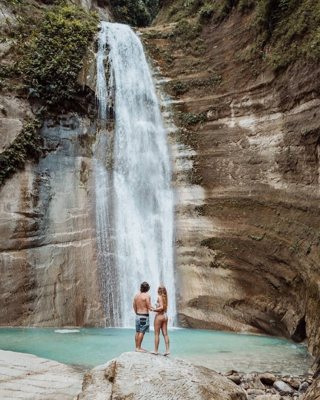 A couple stands hand in hand on a rock ledge in front of a waterfall in Cebu, Philippines. The waterfall cascades into a clear turquoise pool, surrounded by steep rock walls and lush greenery.