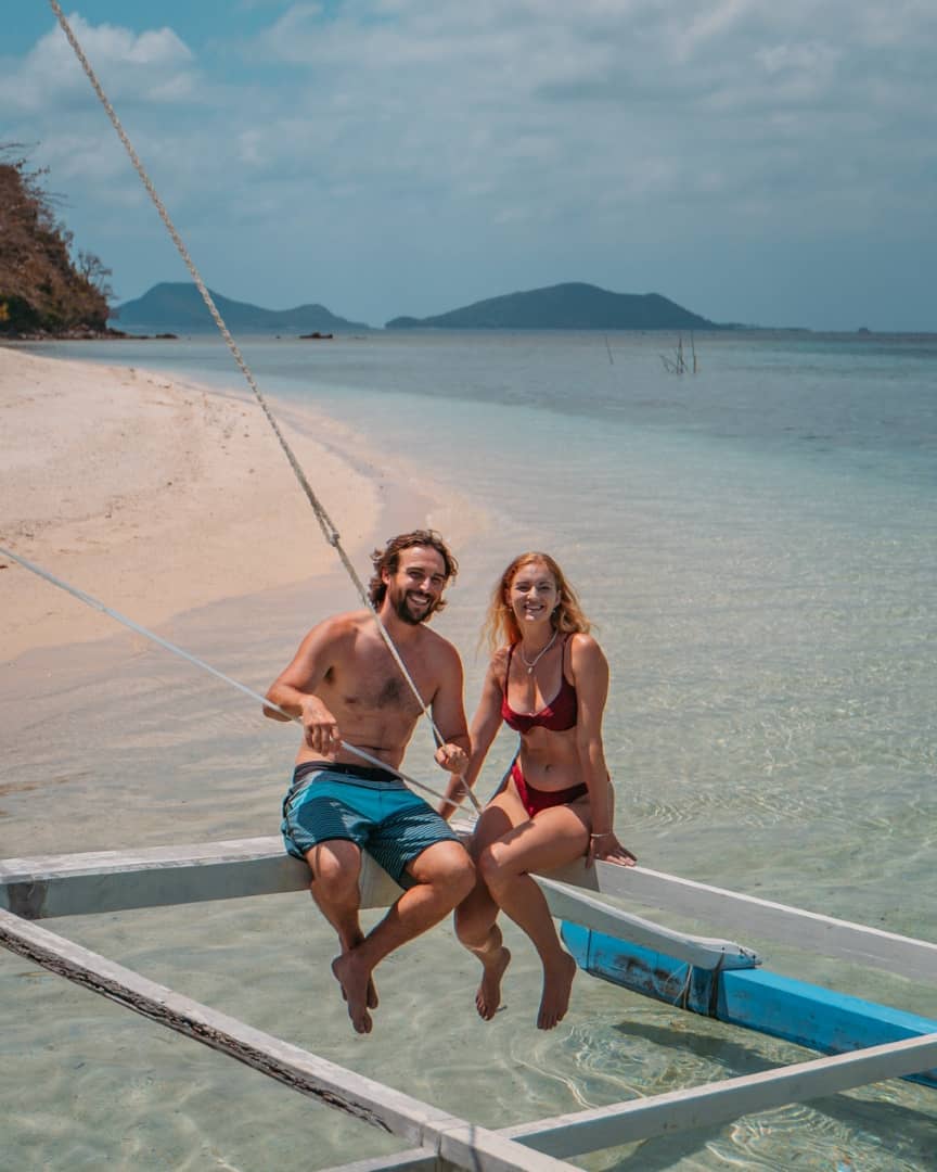 A smiling couple sits on the wooden frame of a traditional Filipino outrigger boat in shallow, crystal-clear water near a sandy beach in Palawan, Philippines. The sea stretches out to distant islands under a partly cloudy sky.
