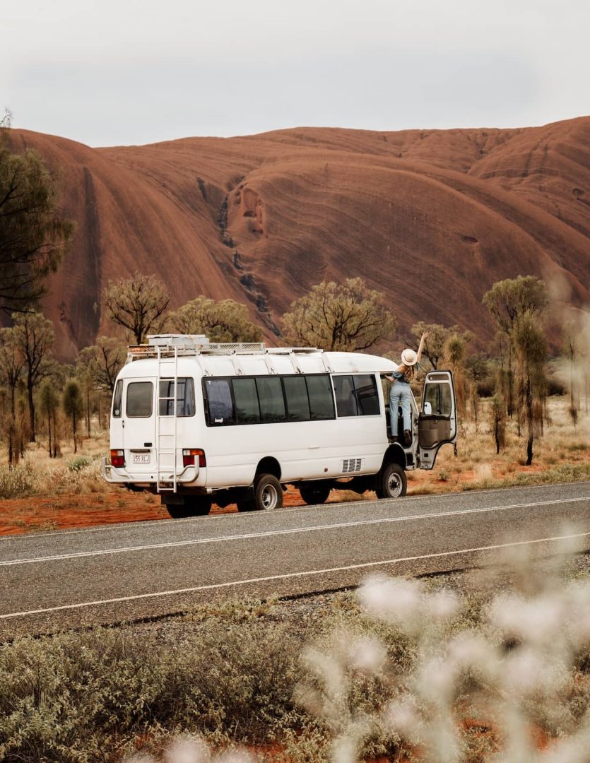 A woman stands in the open doorway of a white van parked by the roadside with Uluru rising in the background during an alice springs to uluru road trip. Native shrubs and muted desert tones frame the scene.