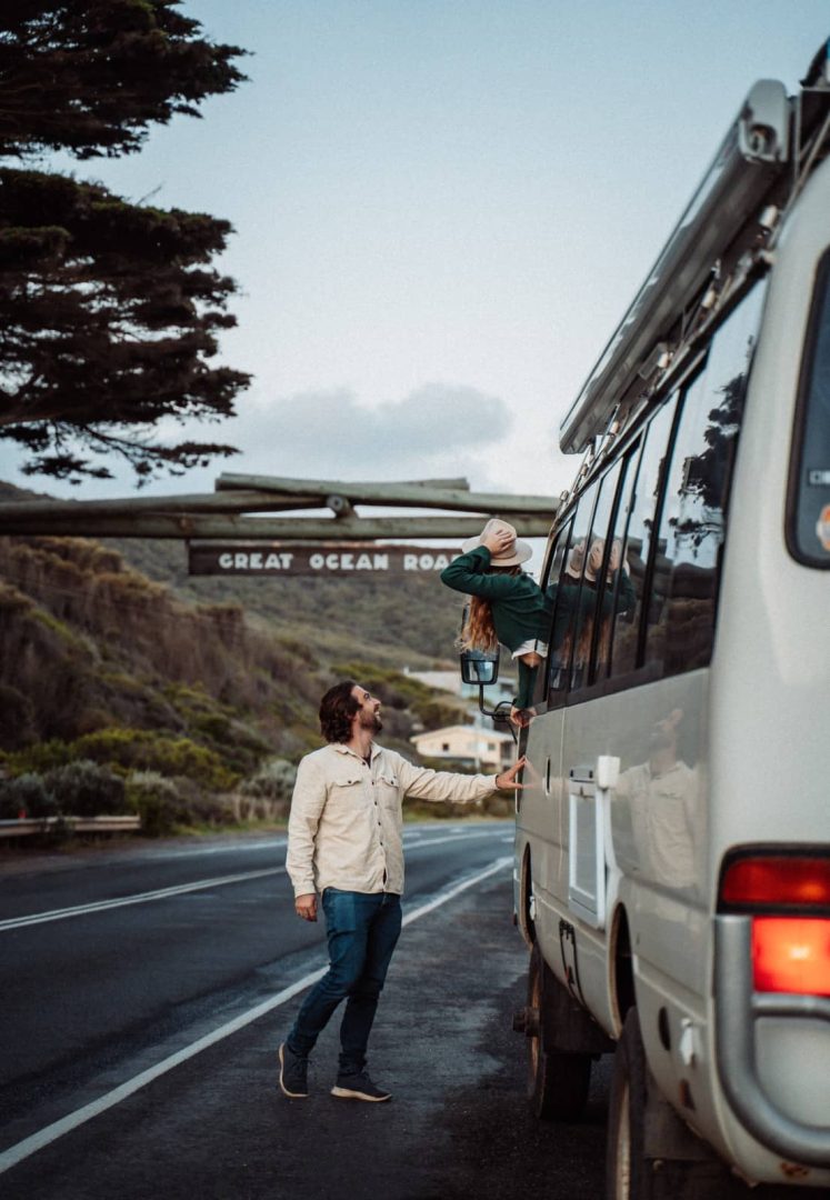 A couple with a campervan stop beneath the iconic Great Ocean Road sign at the start of a Melbourne to Perth road trip, with lush cliffs in the background.