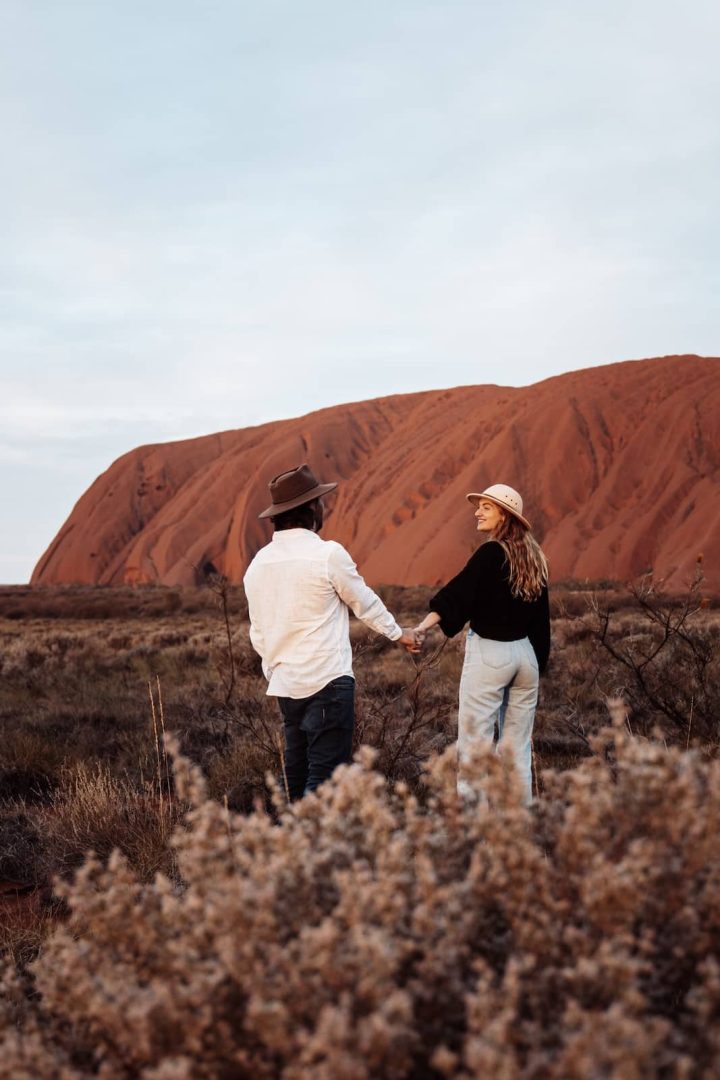 A couple walks hand-in-hand towards Uluru across dry scrubland during their red centre way journey. The massive red rock dominates the background under a pale morning sky.