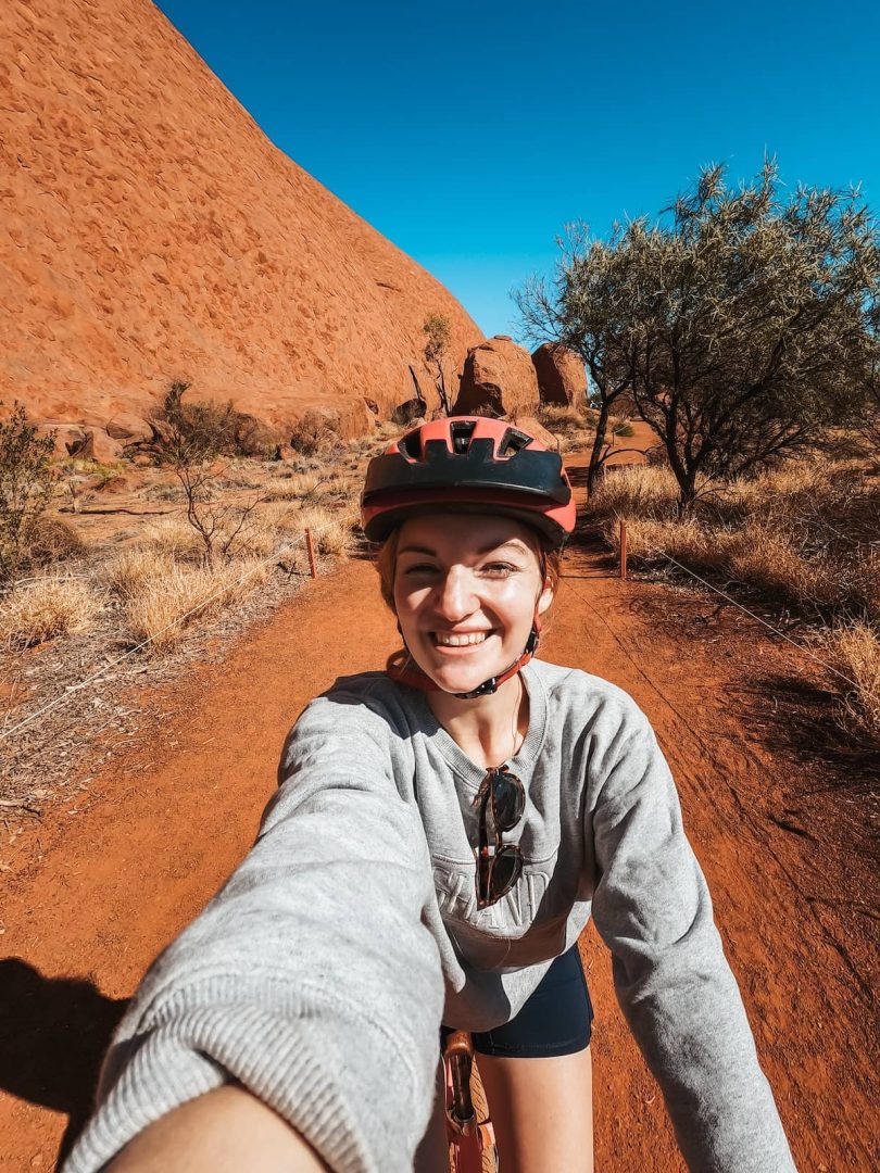 A woman in a helmet smiles while taking a selfie on a bike ride around the base of Uluru during her alice springs to uluru road trip. The deep red earth path and massive rock are visible behind her.