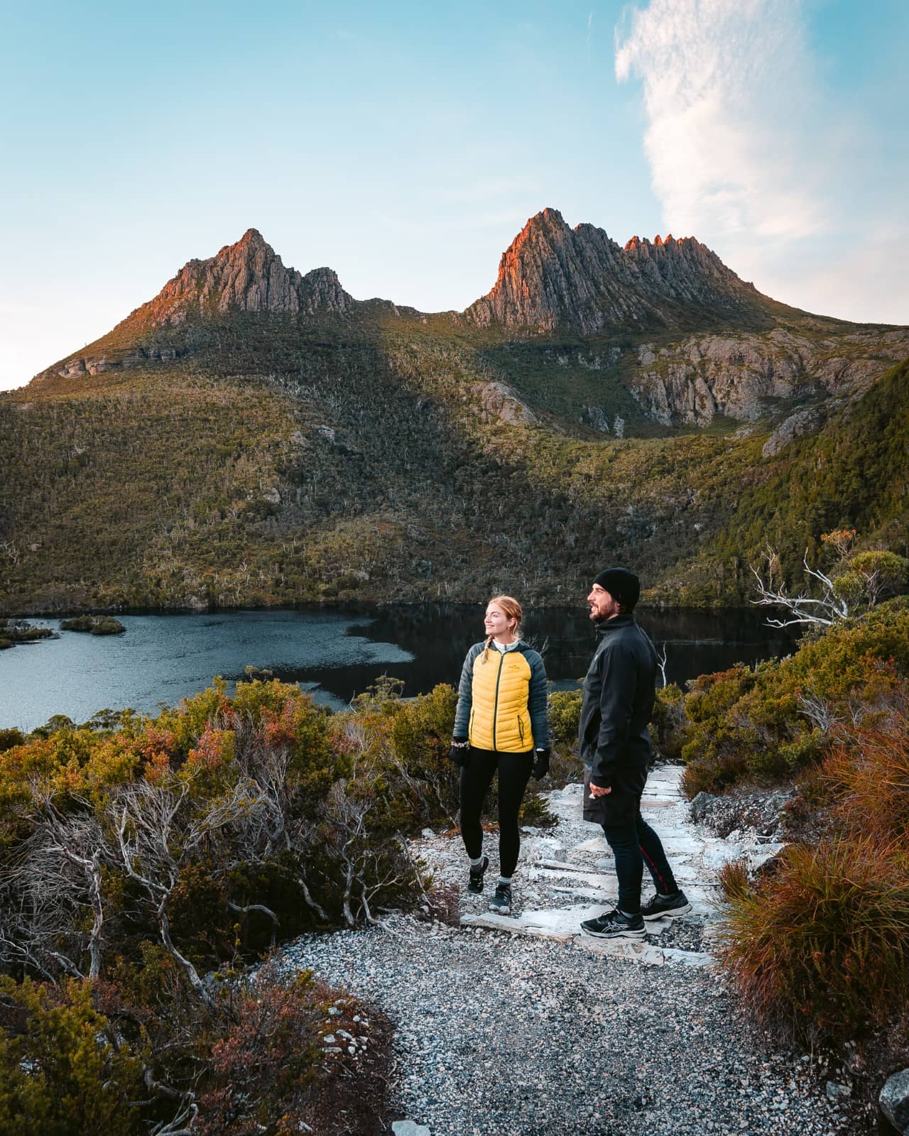 Two hikers on a gravel track overlooking Dove Lake with Cradle Mountain’s rugged peaks behind them at golden hour, a must-see stop when exploring things to do in Tasmania’s national parks.