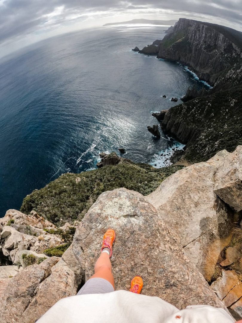 A hiker’s feet in orange shoes stand at the edge of a rocky cliff high above the sea, capturing the vertigo-inducing heights on the Three Capes Track for free.