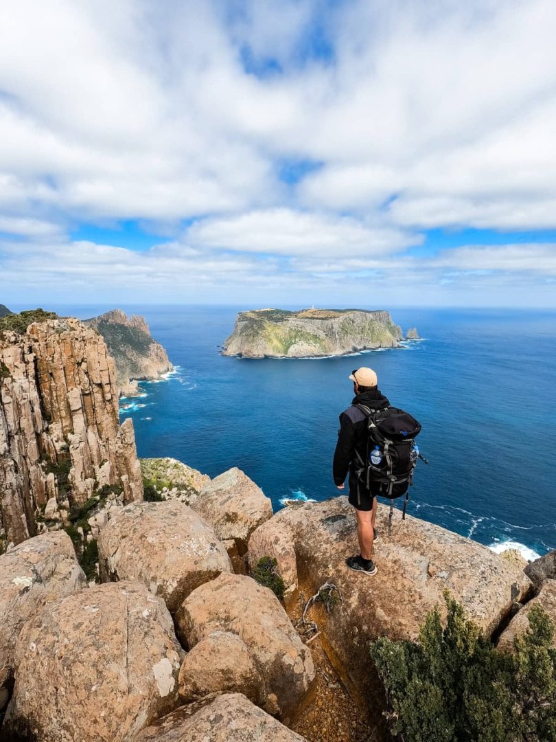 Solo hiker with a backpack looking out over Tasman Island from a cliff’s edge on a clear day, after doing the Three Capes Track, capturing the adventurous side of things to do in Tasmania.