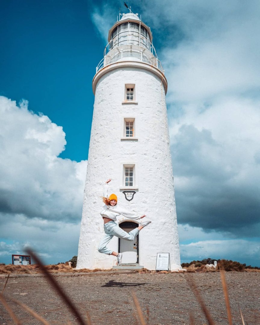 Person mid-jump in front of Cape Bruny lighthouse under a dramatic sky, one of the quirky and photogenic things to do in Tasmania’s coastal areas.