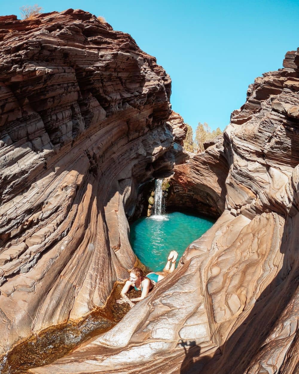 Woman lounges in natural rock formation with small waterfall and turquoise pool, Karijini National Park, under clear blue sky.