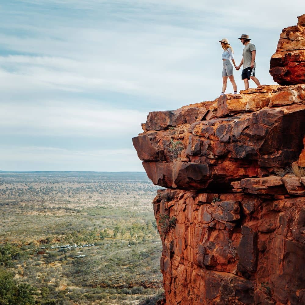 A man and woman holding hands while looking down over the edge of a mountain. Something to see while hiking the very best tracks of the Northern Territory. This is the Giles Track in KIngs Canyon, Alice Springs.