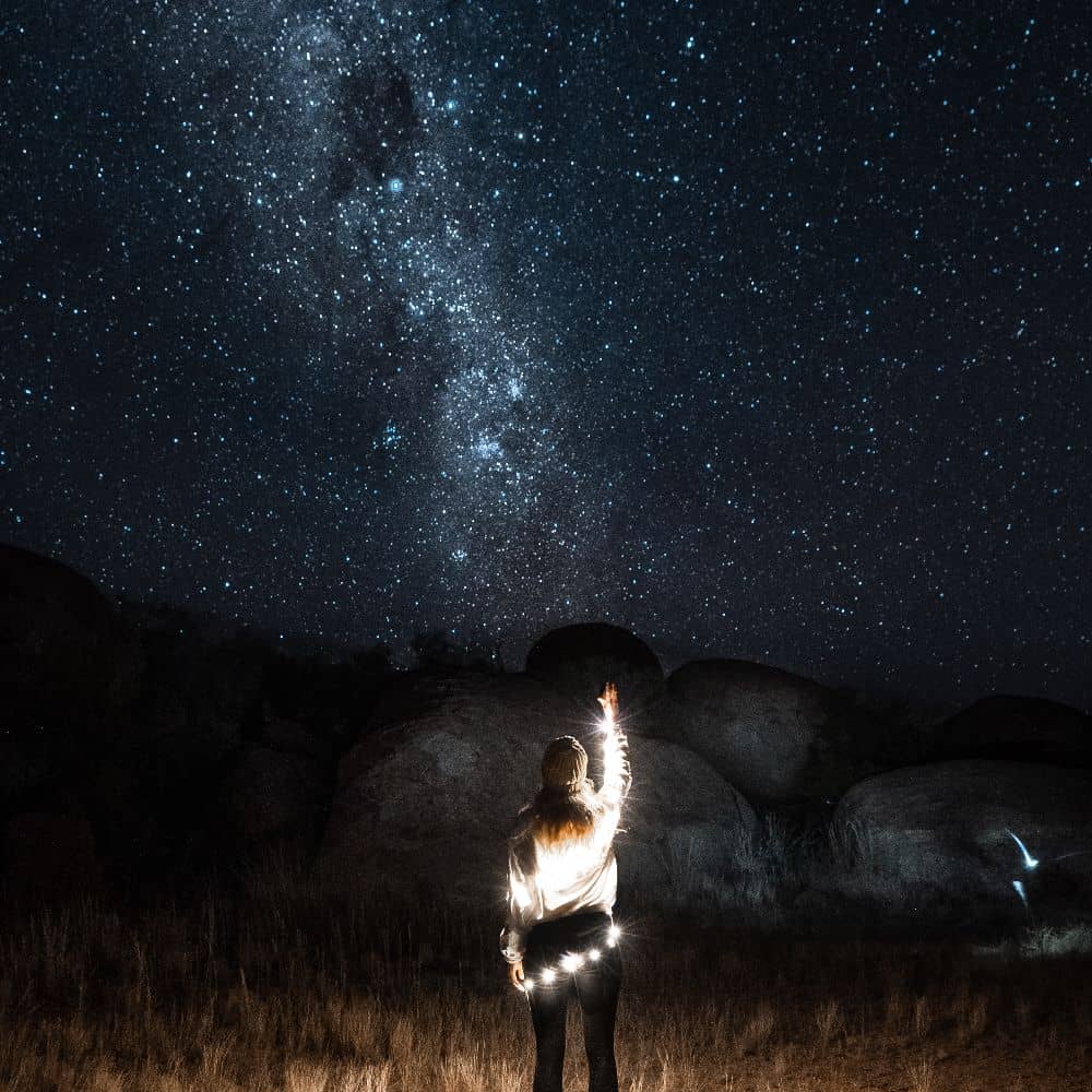 Woman looking up and reaching into the sky, star gazing, with fairy lights wrapped around her. One of the top 15 best sites to see in Alice Springs. Can be found at Alice Springs in the Northern Territory. 