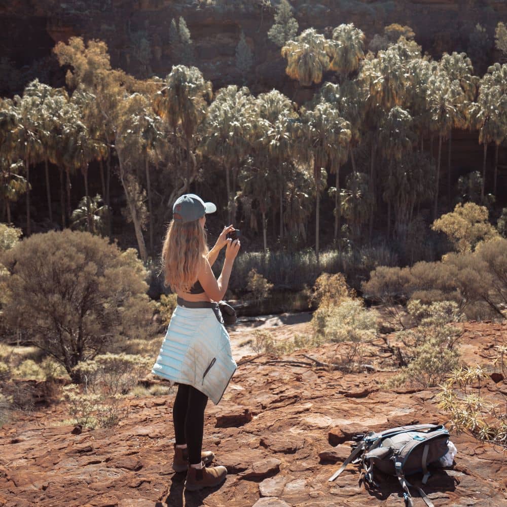 A woman looking over a mountain cliff while holding a camera. One of the top 13 things to do in Alice Springs. Located at the West MacDonnell Ranges in Alice Springs, Northern Territory.