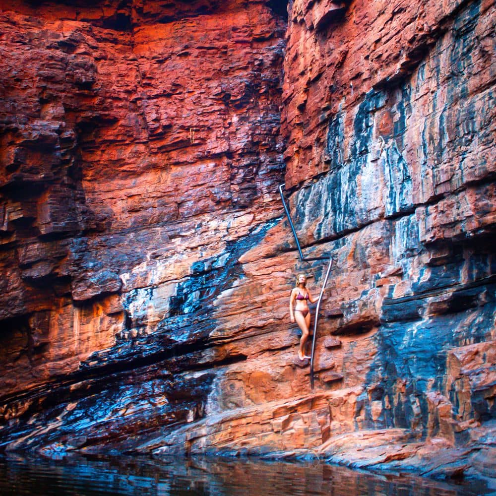 Women looking up into rocks while standing on a rock and holding a handrail above a body of water. Karijini considers it one of the best sites. This is the Handrail Pool located in the Weano Gorge.