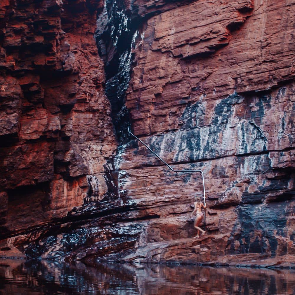 Woman holding a handrail that leads down to a body of water surrounded by red rocks. One of the sites to see in Weano Gorge, Karijini, where you can walk through 2 billion years of history.