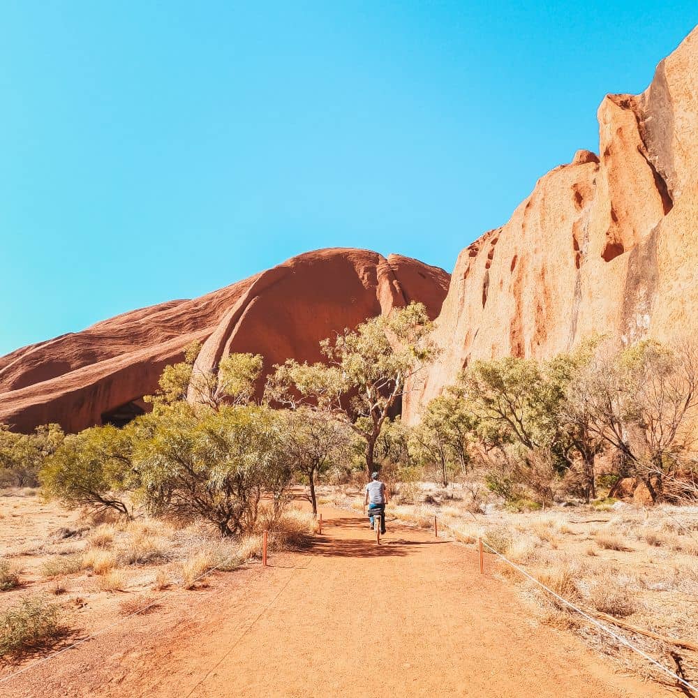 A man riding a bicycle around Uluru under clear skies. Clearly the very best of hiking in the Northern Territory. This popular biking trail can be found at Uluru in Alice Springs.
