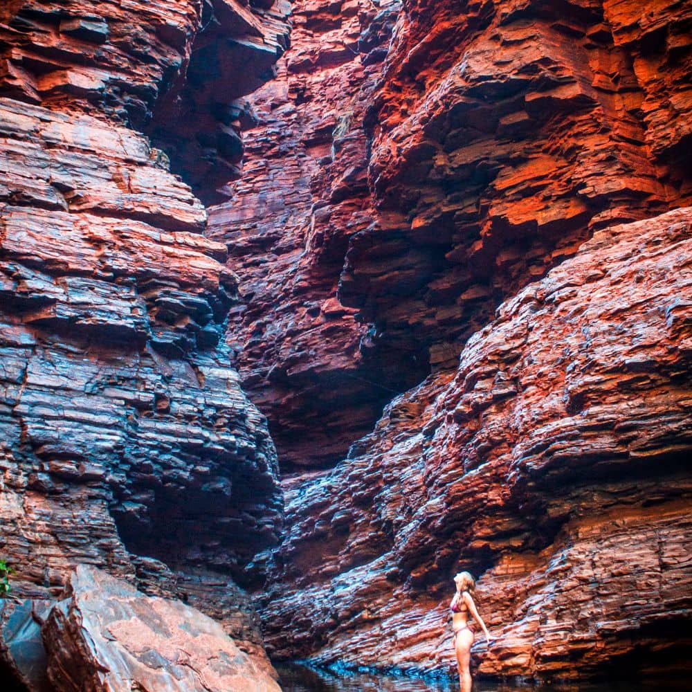 Woman standing in water while looking up into the red rocks she is surrounded by. One of the best things to do in Weano Gorge, Karijini, the place where you can walk through years of history. She is at the Handrail pool in WA.