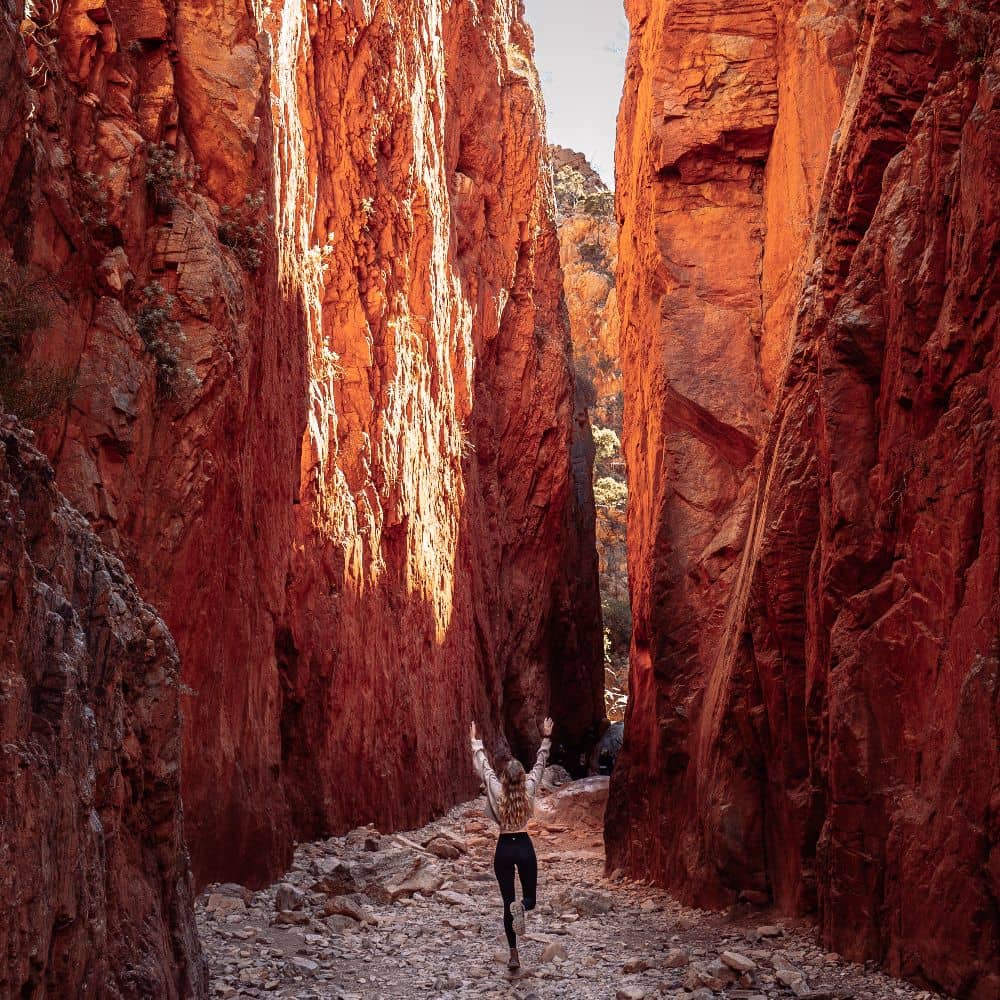 Woman running through a gap in a rock formation with hands in the air. Many locals say it is one of the best things to see in Alice Springs. She is at Standley Chasm.