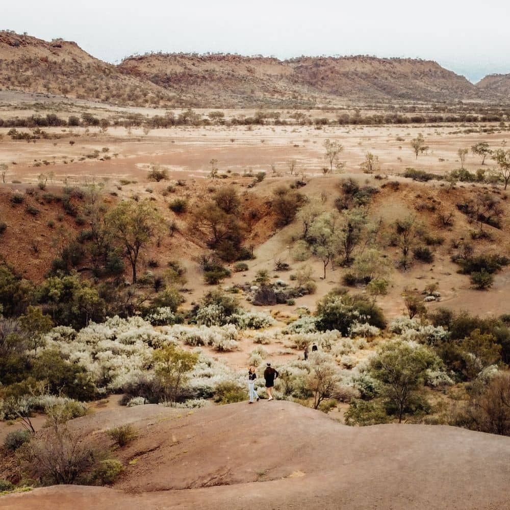 A man and woman standing on a massive rock while looking out at meteor craters. Something cool to see in Alice Springs. This is located at the Gosses Bluff crater site in Alice Springs.
