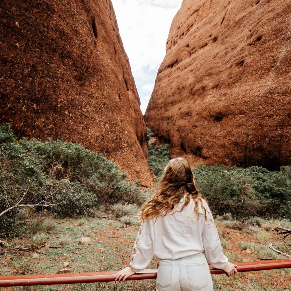 Woman standing on bridge with wind blowing hair and looking out at two rock formations. Something to see when hiking the very best in the NT. This spot is located at the Kata Tjuta National Park in Alice Springs.