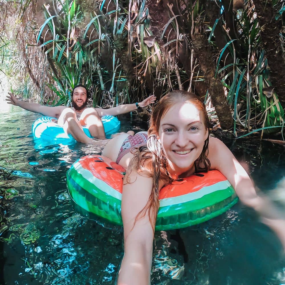 A man and woman sitting on inflatable rings while floating down a river, smiling and taking a selfie. Many tourists say the Katherine Hot Springs are both unforgettable and able to energize the mind. This is the swimming hole at Katherine Springs.