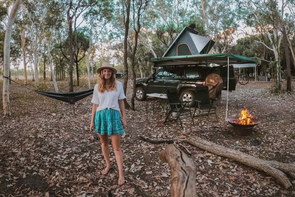 A woman stands at a bush campsite at Mt Elizabeth Station, with a 4WD, rooftop tent, campfire, chairs and hammock set up beneath tall gum trees—a classic overnight experience on a gibb river road itinerary.
