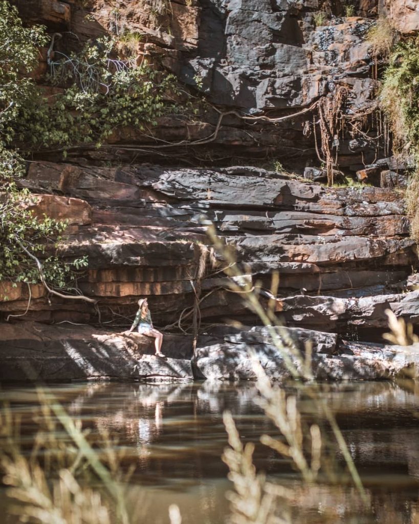 A woman sits on a rock ledge beside the calm water at Adcock Gorge, framed by dark stone cliffs and hanging vines—one of the quieter, shaded swimming spots on a typical gibb river road itinerary.