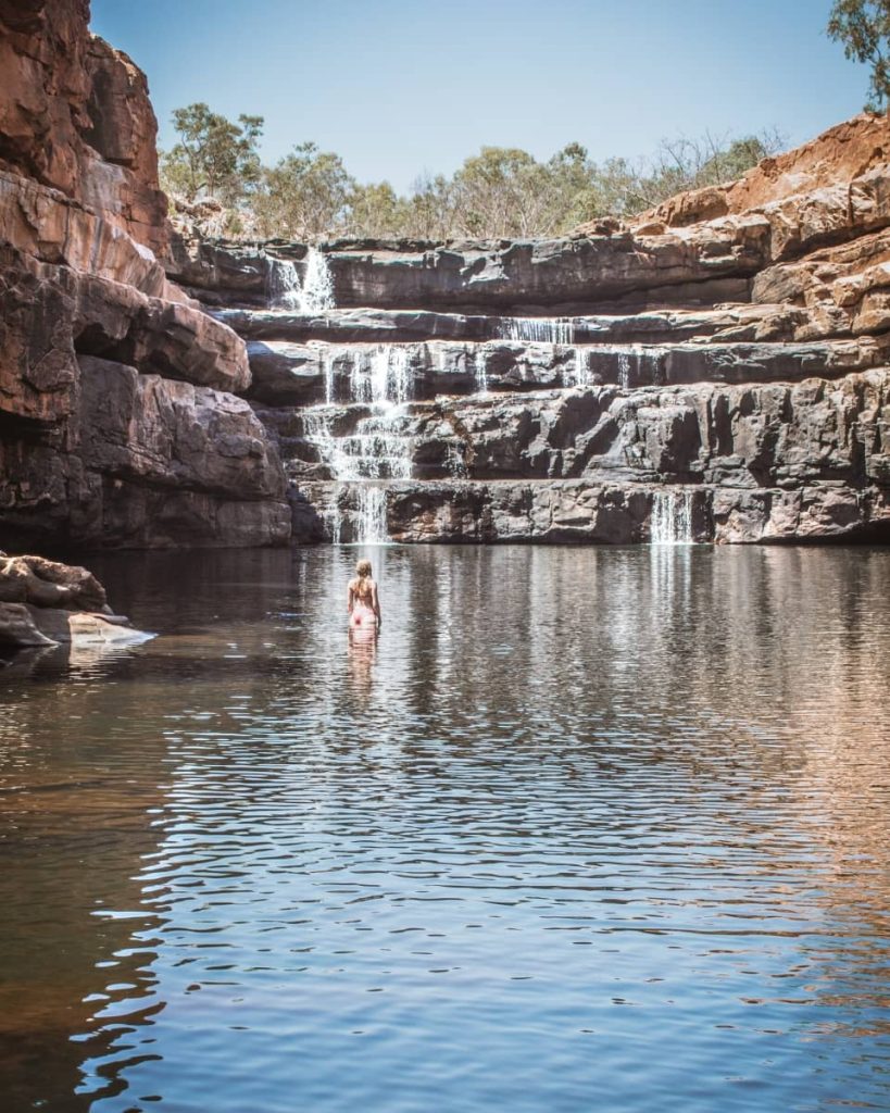 A woman stands waist-deep in the still plunge pool at the base of Bell Gorge, facing the tiered waterfall cascading over layered rock formations—one of the most iconic swimming spots on a gibb river road itinerary.