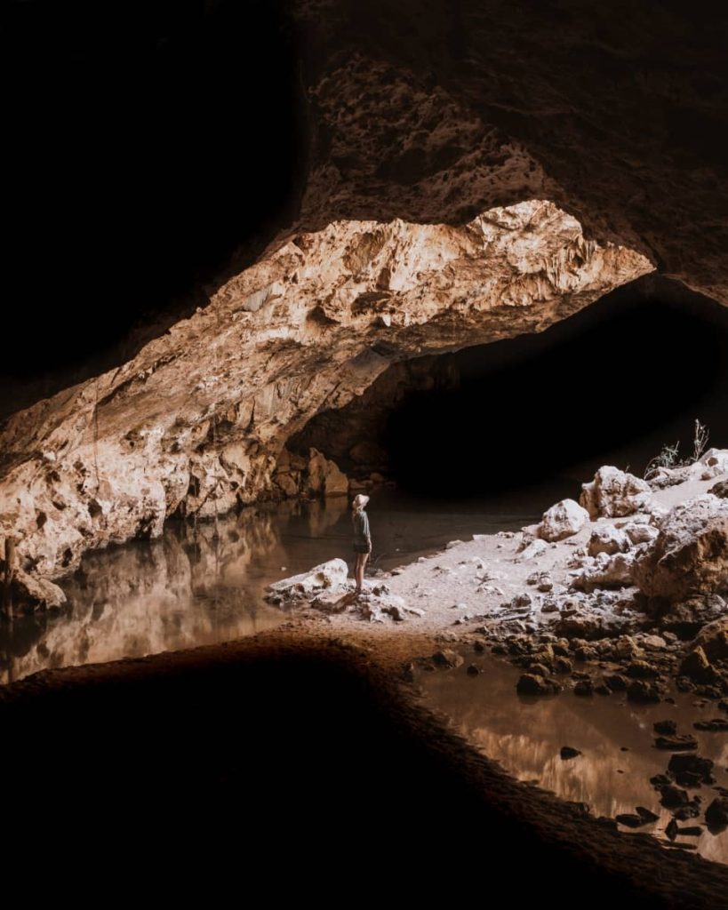 A person stands on a sandy patch inside Tunnel Creek, gazing up at the natural rock arch lit by filtered light—this atmospheric cave system is a fascinating stop on a gibb river road itinerary.