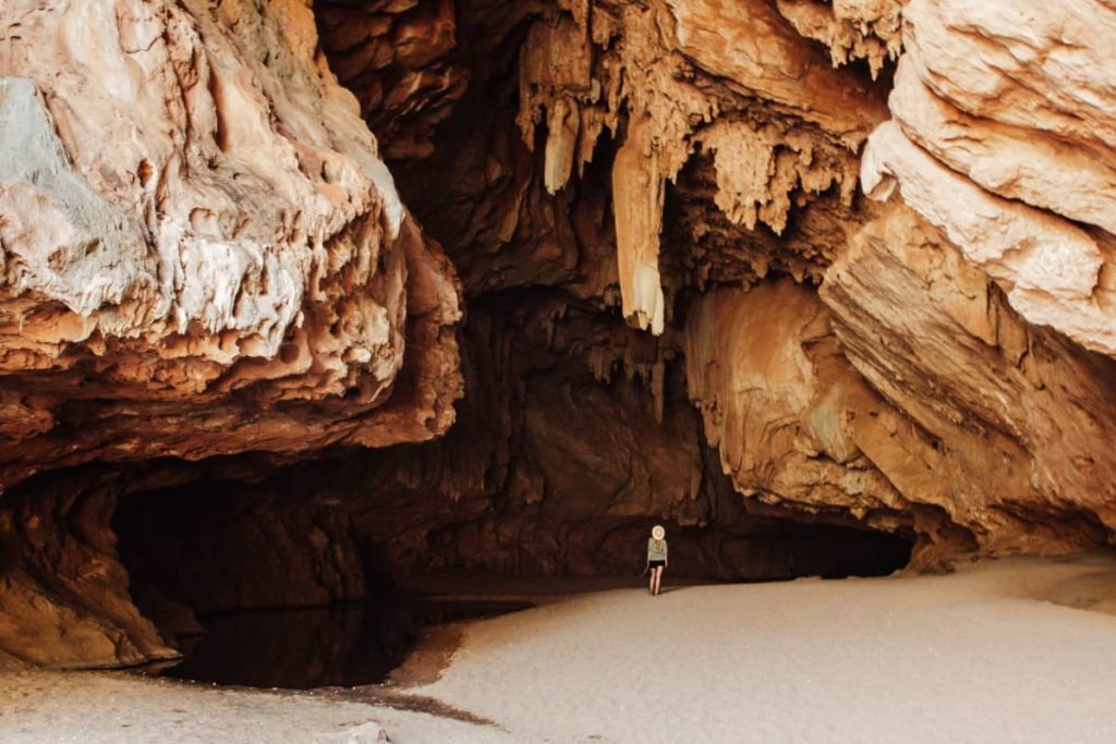 A woman stands on sandy ground beneath the towering rock ceiling of Tunnel Creek, surrounded by dramatic limestone formations and natural textures—an atmospheric highlight of a gibb river road itinerary.