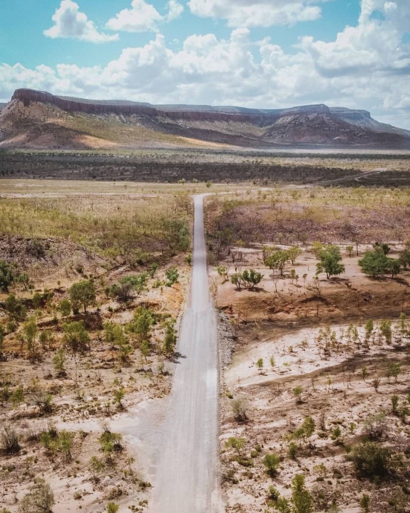 Straight section of gravel road cutting through open terrain near mountain ranges under a blue sky—showing the rugged scenery between Derby and Kununurra on a gibb river road itinerary.