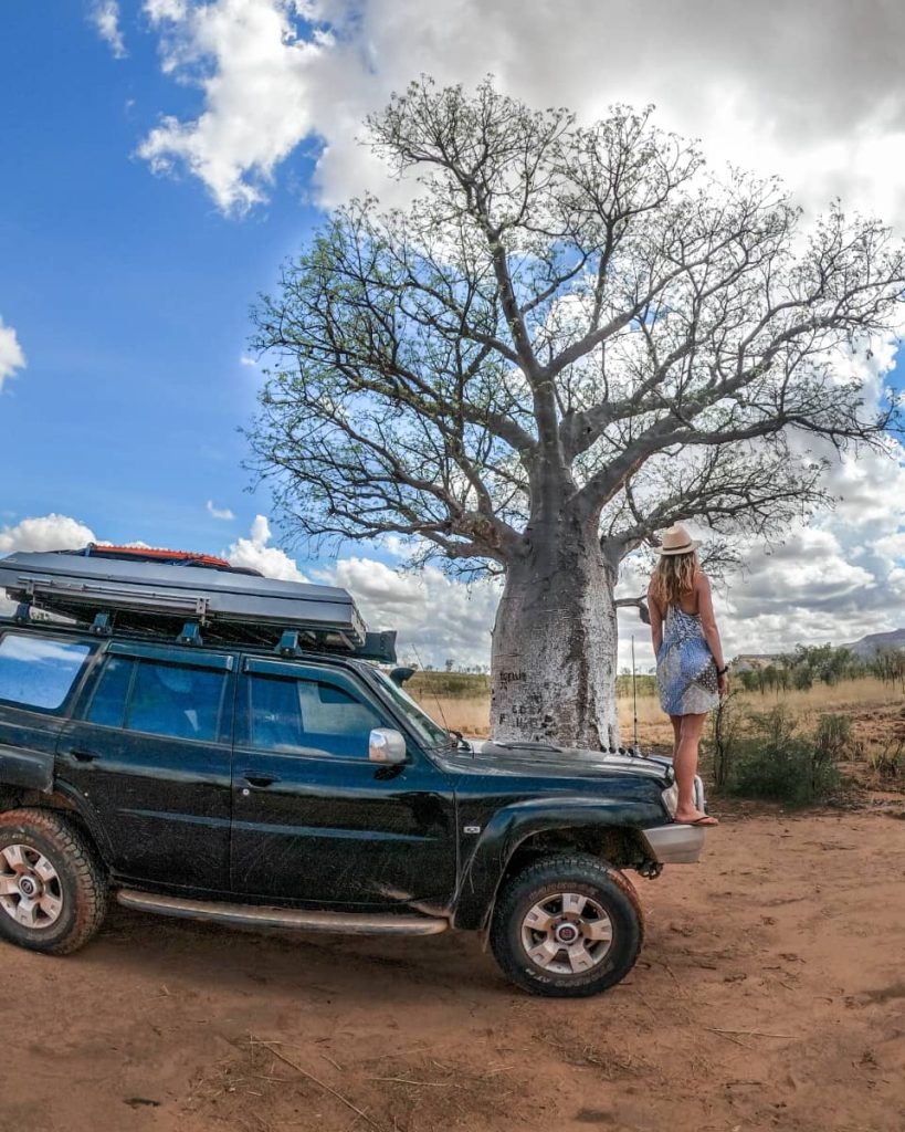 A woman stands on the bonnet of a 4WD parked next to a large boab tree under a partly cloudy sky—capturing a classic outback moment on the gibb river road itinerary.
