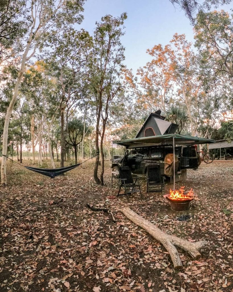Campfire glows beside a 4WD with rooftop tent set up among tall trees, with chairs and hammock strung up—showing a cheap paid camping site on a gibb river road itinerary.