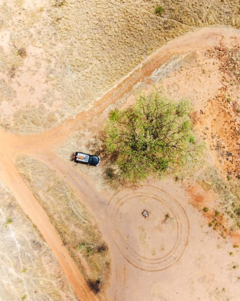 Aerial view of a 4WD parked under a lone green tree beside a dirt track in the Kimberley outback, with tyre marks forming a circle on the red earth—typical of remote camp stops along a gibb river road itinerary.