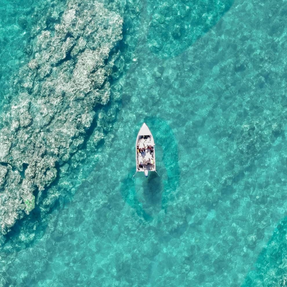 An aerial view of a small boat floating on the crystal-clear turquoise waters of Ningaloo Reef, with the coral formations visible beneath the surface.