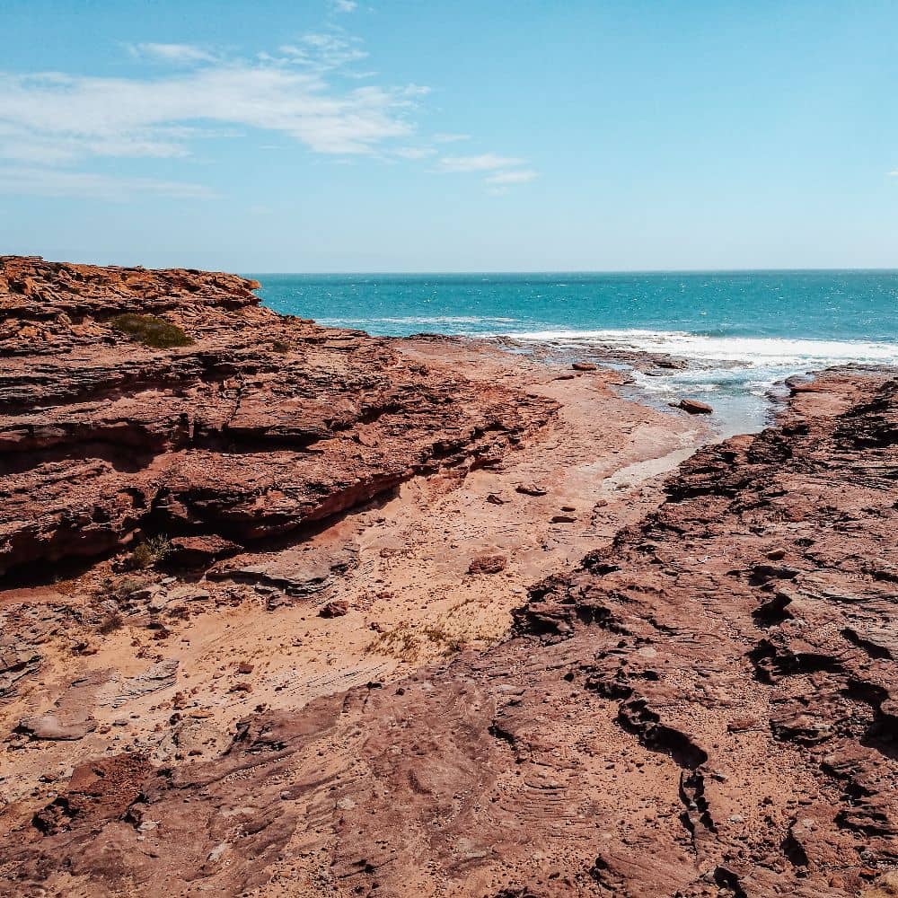 A rugged, reddish-brown coastline at Red Bluff Beach, Kalbarri, meets the bright blue ocean under a clear sky.