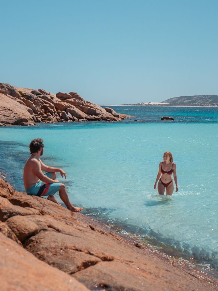 Chris and Bec in Wharton Beach, Chris is sitting on a rock looking at Bec who's walking out the turquoise waters