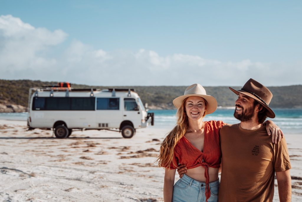 Chris and Beck embrace in front of Salt and Charcoal camper van parked on the sandy shore, with clear blue waters and a sunny sky in the background in Bremer Bay beach Western Australia