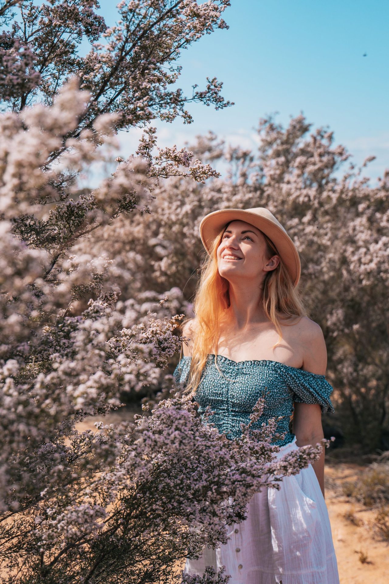 A woman in a straw hat and off-shoulder blue top stands amidst a blooming field of delicate pink wildflowers in Western Australia, looking up at the sky with a bright smile.