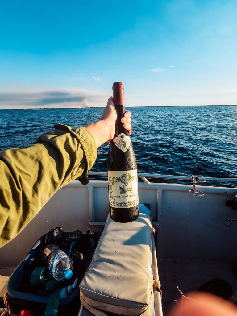 First-person perspective from a boat near Busselton Jetty, Western Australia, holding up a bottle of Albariño de Fefiñanes wine against the serene blue ocean and clear sky.