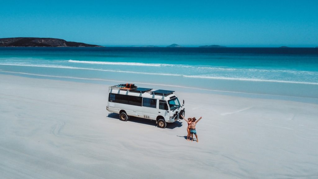 Aerial shot of the Salt and Charcoal van and two people on the white sands of Cape Le Grand beach near Esperance, with the turquoise ocean and distant islands in the background.