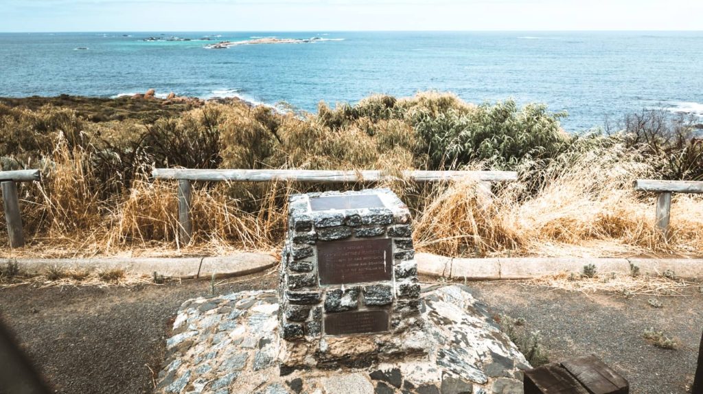 A commemorative plaque of Captain Matthew Flinders on a stone pedestal overlooking the scenic coastline near Augusta, Western Australia, set against a backdrop of ocean and wild coastal vegetation.