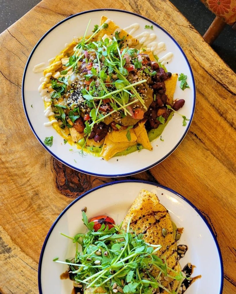 Two dishes on a wooden table: one with nachos, black beans, avocado, and greens, the other with a crisp tortilla and salad, both drizzled with sauce.