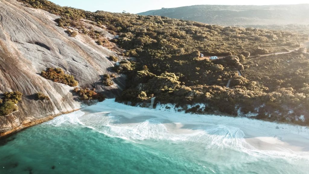 An aerial view of the serene beauty of Misery Beach in Albany, Western Australia, with its turquoise waters gently lapping against a white sandy shore, bordered by large smooth granite rocks and dense green vegetation, with a couple of vehicles parked amidst the trees, highlighting the secluded nature of this coastal paradise.
