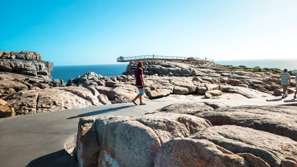 Visitors walk on a pathway among the rounded, weathered rocks leading to a viewing platform at The Gap in Albany, Western Australia, with the vast ocean extending to the horizon.