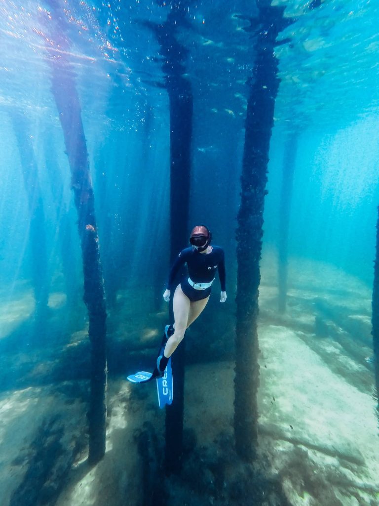 Scuba diver in blue gear hovering peacefully in the clear, turquoise waters of Busselton Jetty's Underwater Observatory, Western Australia, surrounded by vibrant marine life and colorful coral encrusted pillars.
